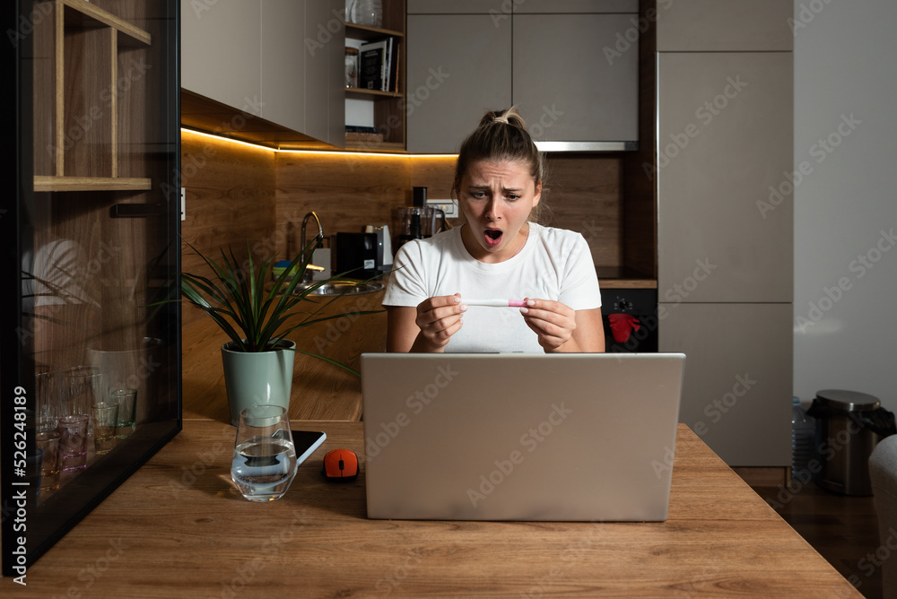 Young woman surprised negative facial reactions on positive result on pregnancy test. Young girl talking on video call on laptop computer with her mother complaining that she is pregnant. Unwanted Young woman surprised negative facial reactions on positive result on pregnancy test. Young girl talking on video call on laptop computer with her mother complaining that she is pregnant. Unwanted