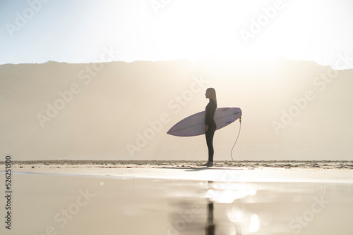 Beautiful surfer girl at the beach standing with her surfboard at sunset time. Female surfer
