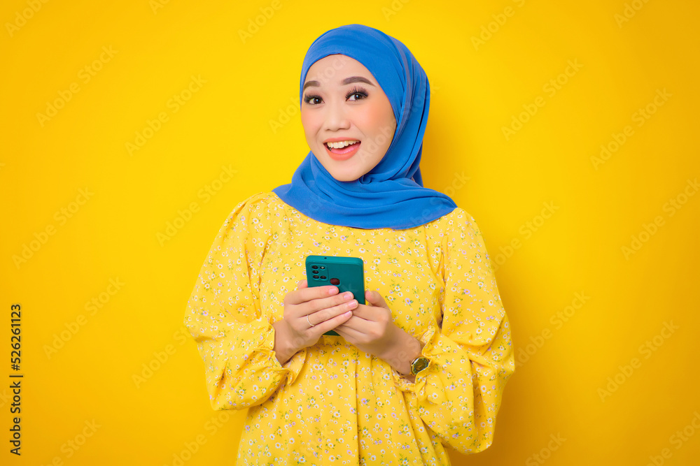 Cheerful young Asian woman in casual dress holding mobile phone and looking at camera with happy expression isolated on yellow background Cheerful young Asian woman in casual dress holding mobile phone and looking at camera with happy expression isolated on yellow background