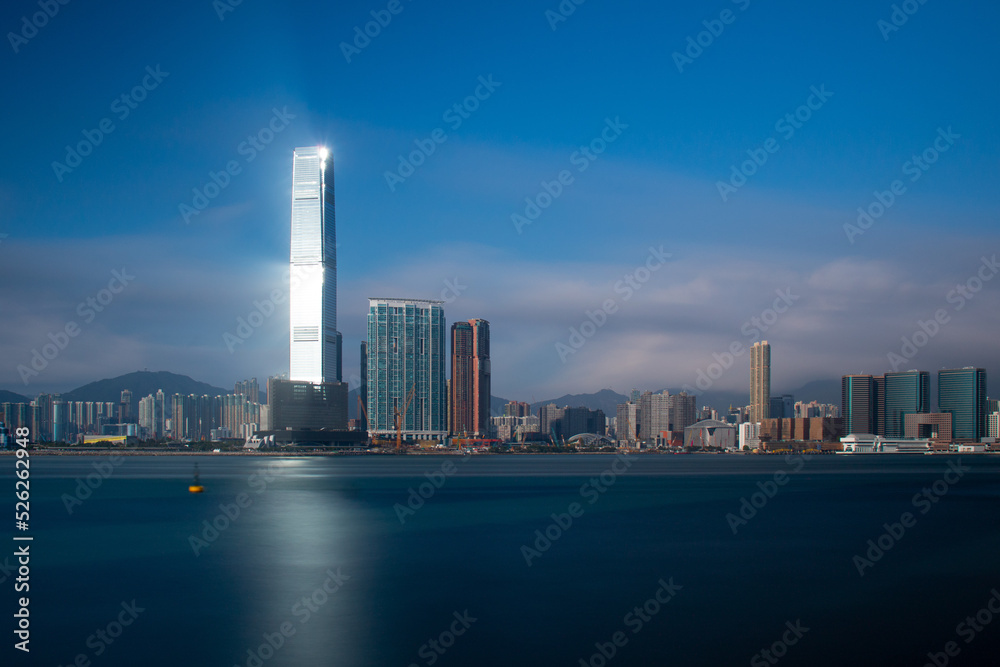 Lighthouse.  The sun reflects off a skyscraper on Kowloon, Hong Kong casting light over the blue sea.  Long exposure gives a silky smooth sea.
