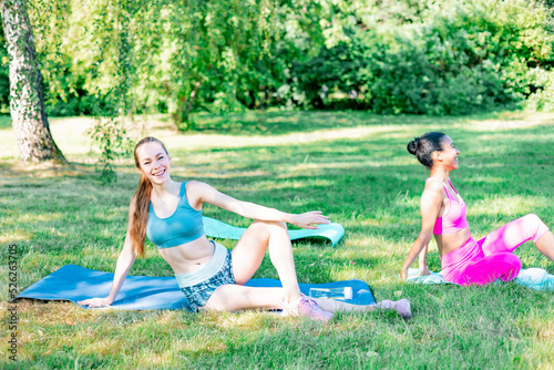 beautiful young multi ethnic women friends do sports fitness or yoga on mat in park on outdoor on sunny summer day, healthy lifestyle, training and stretching in open air together © klavdiyav