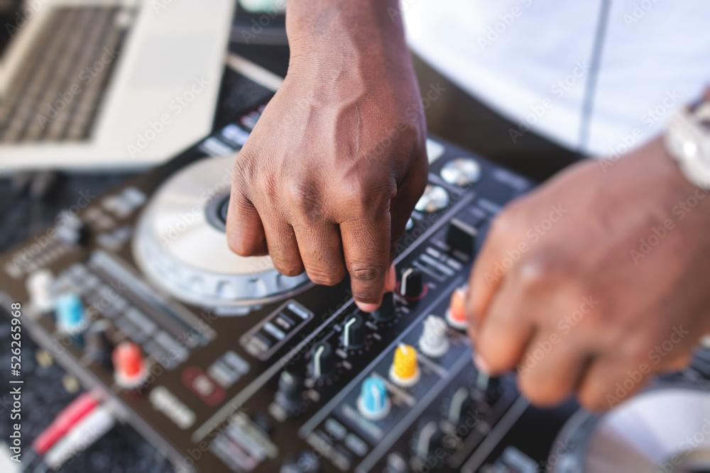 View of Dj mixer and vinyl plate with headphones on a table with african american DJ playing on stage and mixes the track in the background, during summer open air event techno party, hand close up View of Dj mixer and vinyl plate with headphones on a table with african american DJ playing on stage and mixes the track in the background, during summer open air event techno party, hand close up