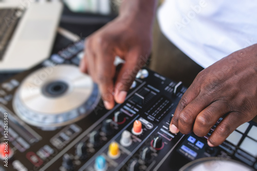 View of Dj mixer and vinyl plate with headphones on a table with african american DJ playing on stage and mixes the track in the background, during summer open air event techno party, hand close up