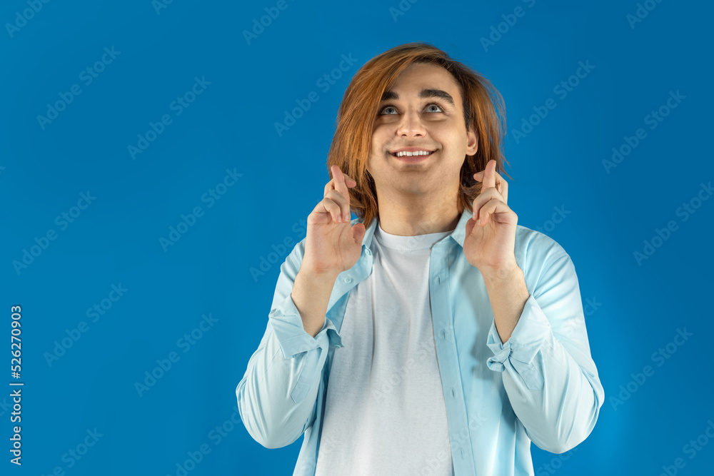 Happy young man Prep Student with long hair. Portrait of a guy keeping fingers crossed on a blue background Happy young man Prep Student with long hair. Portrait of a guy keeping fingers crossed on a blue background