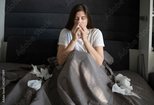 Woman lying in bed holds napkin tissue near her face. Lots of scattered handkerchiefs. Woman holds napkin near nose, sneezes, suffers from runny nose. Concept: coronovirus, flu epidemic