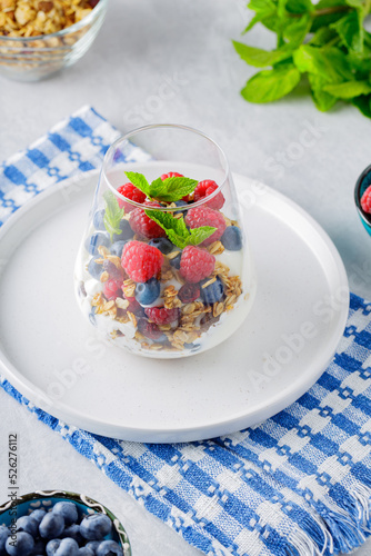 Glass with granola, yogurt, fresh raspberries, blueberries and mint. High protein and diet breakfast on a white-blue napkin. Healthy food concept