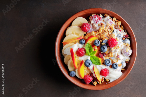 Granola breakfast with yogurt, blueberry, raspberry, banana and peach. Healthy breakfast with yogurt, baked granola and fruit on a clay plate. Top view. Copy space