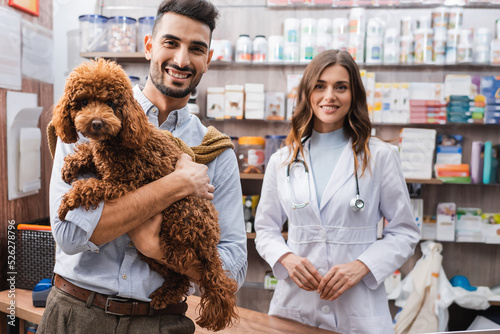 Smiling arbian man holding poodle near veterinarian in pet shop photo