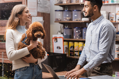 Side view of smiling woman holding poodle near arabian seller in pet shop