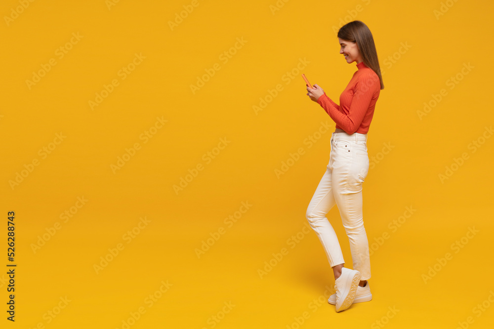 Side view of woman standing on yellow copy space background chatting online using phone - obrazy, fototapety, plakaty 
