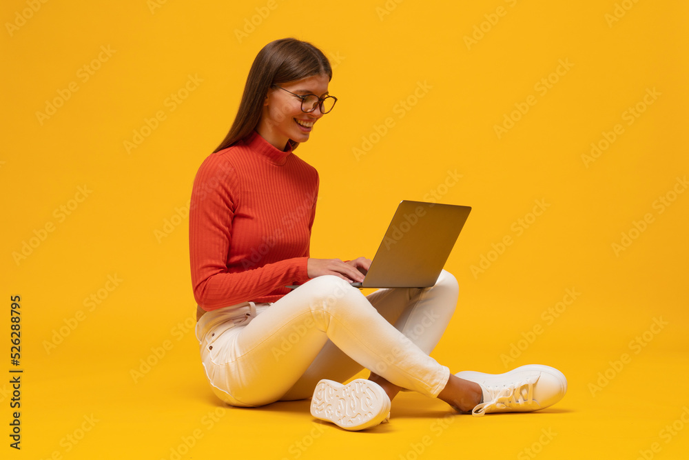 Side view portrait of woman browsing social media on laptop sitting on floor on yellow background Side view portrait of woman browsing social media on laptop sitting on floor on yellow background
