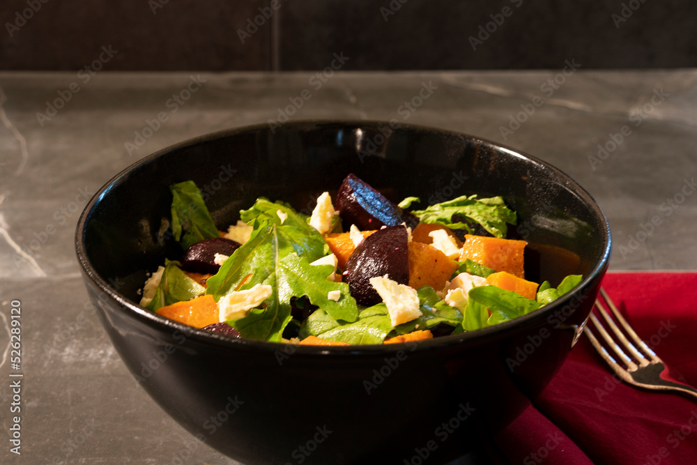 A salad made of roasted pumpkin, beetroot and feta cheese served in a black bowl against a textured grey background.