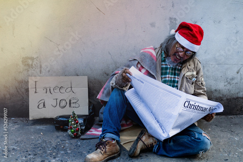 Fotografie A homeless man, an old Asian man wearing a red Christmas hat, is sitting on the ground reading a newspaper, Look for jobs from newspapers