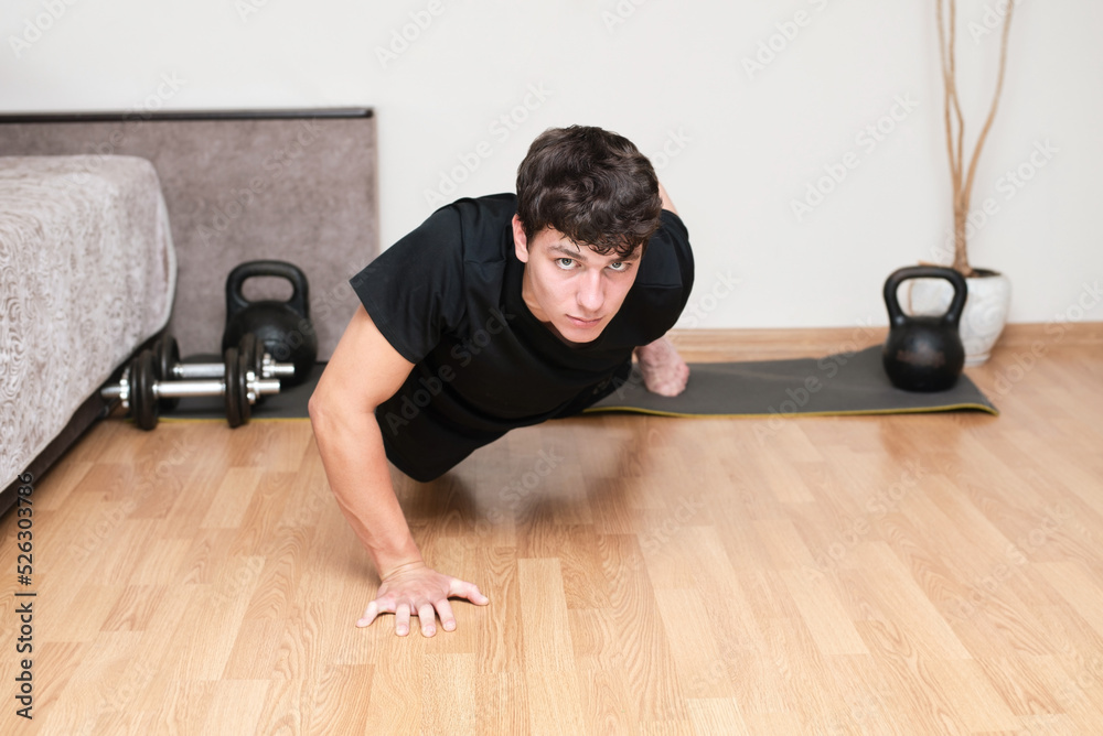 Young teenage guy exercising at home, has an athletic build, does push-ups from the floor on one hand, behind him is a gym mat and dumbbells with kettlebells