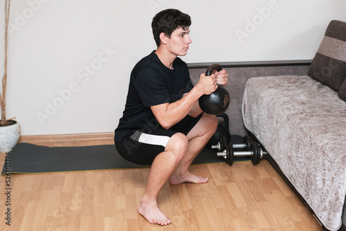 Young man does not always have time to go to the gym and often works out at home, now squats with a heavy kettlebell, sports concept, he has a focused but happy face