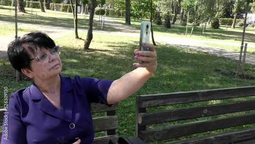 A woman sits on a bench and takes a selfie.
On a summer day, a woman sits on a park bench and takes a selfie with her phone.
