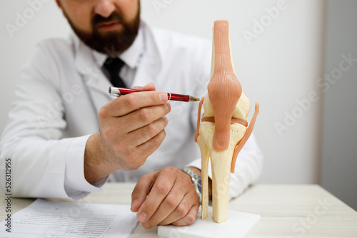 Male doctor shows model of human artificial knee joint in medical office photo