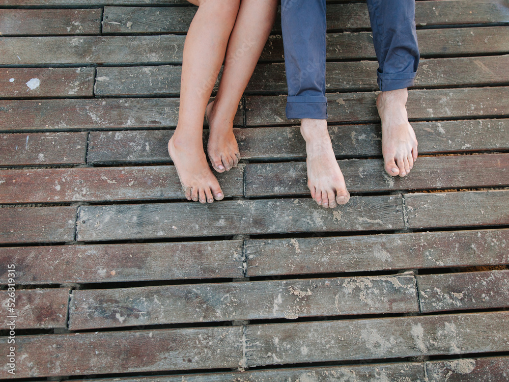 A couple is sitting on a wooden pier. The girl has bare legs, the guy is in blue pants, and his feet are on the beach sand.