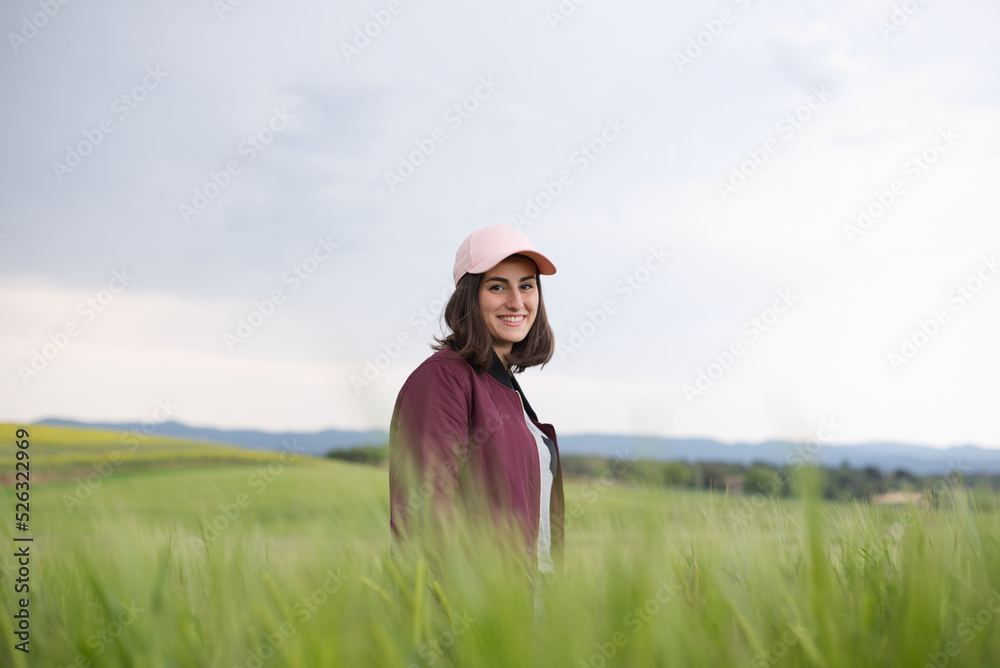 Side view of smiling woman with brown hair and a pink cap looking at the camera standing in a beautiful green wheat field. Side view of smiling woman with brown hair and a pink cap looking at the camera standing in a beautiful green wheat field.