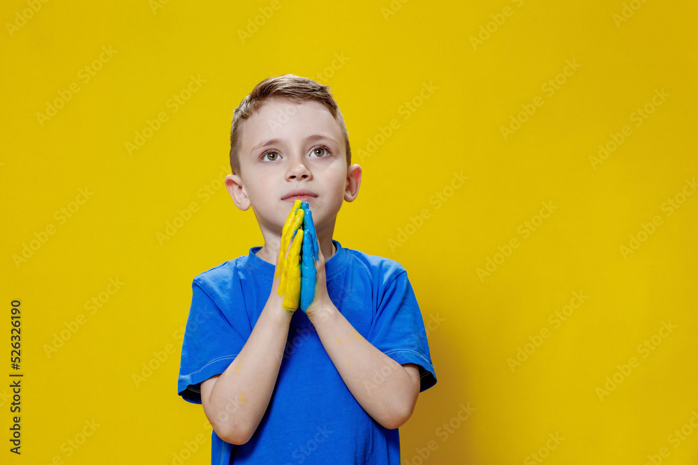Ukrainian boy prays for Ukraine. Children against war. A boy in a blue T-shirt on a yellow background folded his hands painted in the Ukrainian flag for prayer Ukrainian boy prays for Ukraine. Children against war. A boy in a blue T-shirt on a yellow background folded his hands painted in the Ukrainian flag for prayer