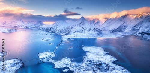 Aerial view of sea, snowy islands, mountains, road, sky with pink clouds at sunset in winter. Lofoten islands, Norway. Landscape with rocks in snow, water at dusk. Top view from above. Nature