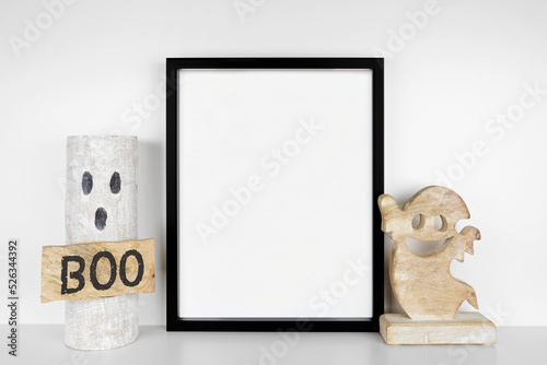 Halloween mock up. Black frame on a white shelf with rustic wood ghost decor. Portrait frame against a white wall. Copy space. #526344392