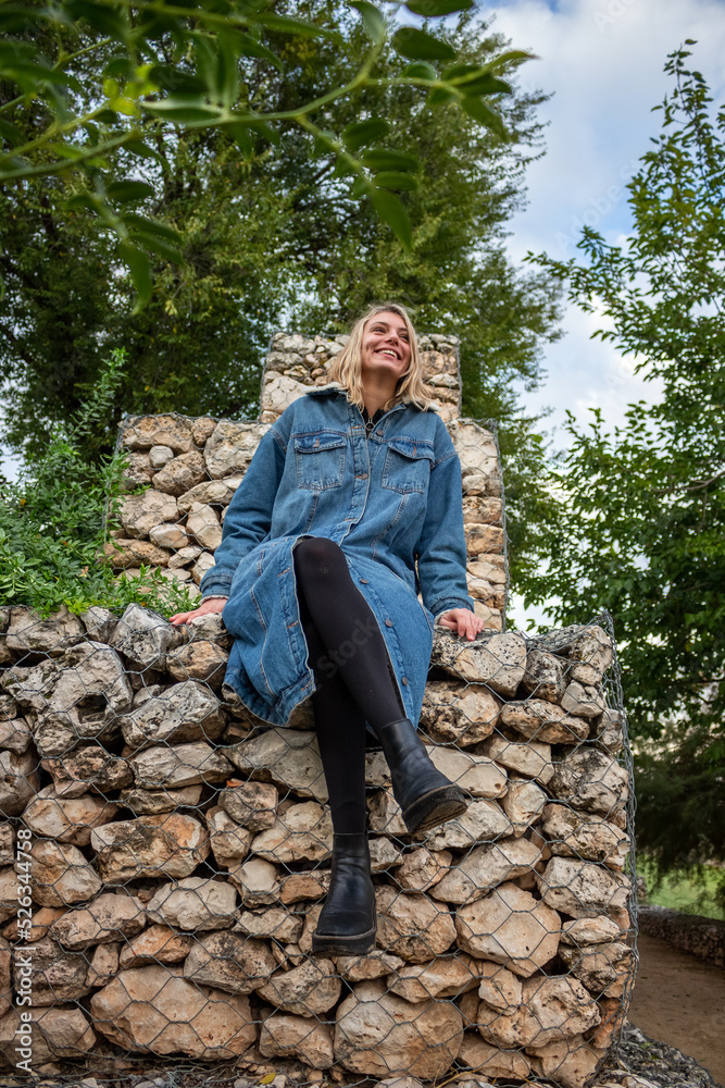 pretty blonde woman sitting on a rock in a green park. She is happy, smiling and has a blue coat pretty blonde woman sitting on a rock in a green park. She is happy, smiling and has a blue coat