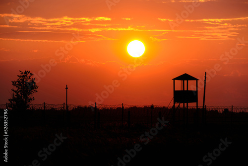 Fence with barbed wire and an old watchtower background bright sun and scarlet sunset.