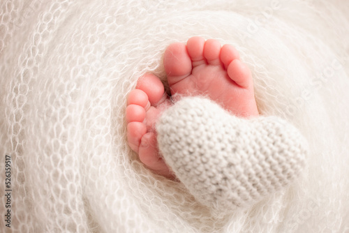 Knitted white heart in the legs of a baby. Soft feet of a new born in a white wool blanket. Close-up of toes  heels and feet of a newborn. Macro photography the tiny foot of a newborn baby.