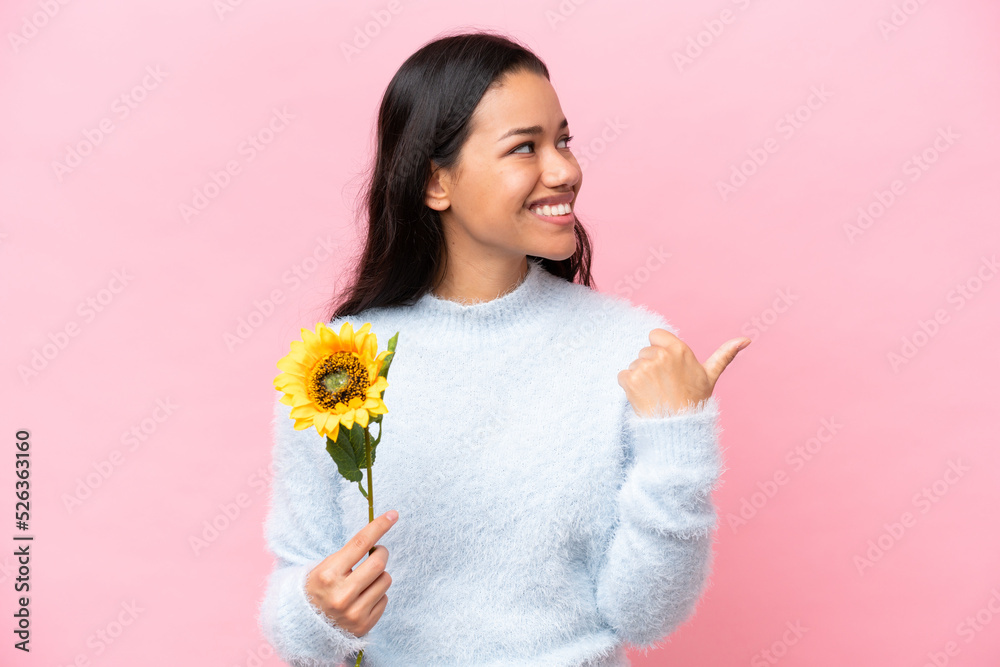 Young Colombian woman holding sunflower isolated on pink background pointing to the side to present a product