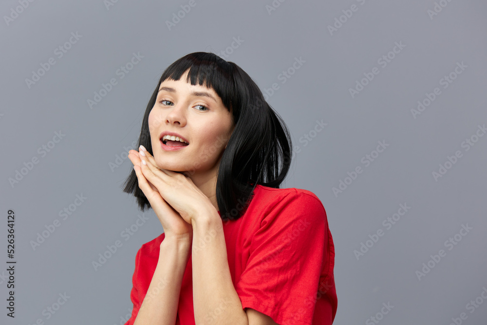 Fototapeta premium a sweet, gentle, sophisticated, relaxed woman with black hair stands in a red T-shirt on a gray background and holds her hands near her face