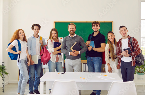 Group portrait of friendly male teacher and his college or high school students. Smiling teacher and students with backpacks on shoulders stand in row on background of blackboard and smile at camera.