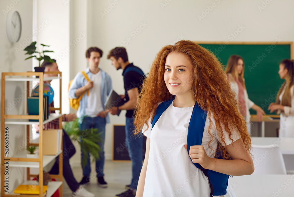 High school, university or college education. Portrait of cute beautiful red-haired girl student in classroom against background of her classmates. Girl with backpack on shoulders is looking at camera