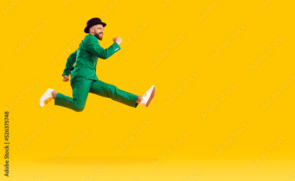 Happy cheerful young man wearing a green suit and a black hat hurrying to a St Patrick's Day party, running fast and jumping high in the air isolated on a bright yellow color copy space background
