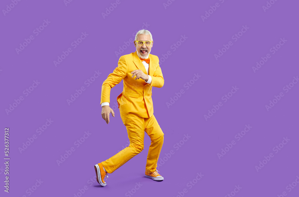 Active life and leisure of older people. Cheerful trendy, eccentric and energetic senior man funny dancing isolated on purple background. Full length of funny male retiree in yellow suit and sneakers. Active life and leisure of older people. Cheerful trendy, eccentric and energetic senior man funny dancing isolated on purple background. Full length of funny male retiree in yellow suit and sneakers.