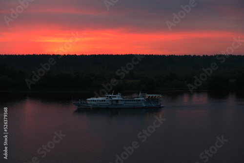 A white steamer floats down the river at night under a colorful red sunset sky