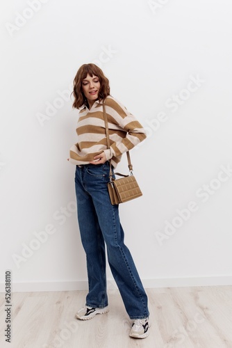 a full-length photo of stylish woman standing on a light background in a striped sweater and blue jeans, posing relaxed with a beige bag worn over her shoulder, adjusting the sweater and looking away