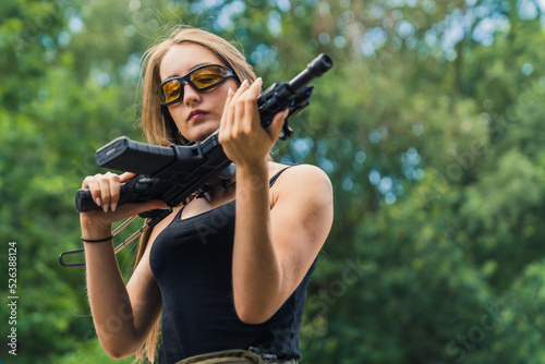 Outdoor shooting range concept. Young beautiful long-haired caucasian girl in a black tank top looking up close at a black rifle. Blurred trees in the background. Medium shot. High quality photo photo