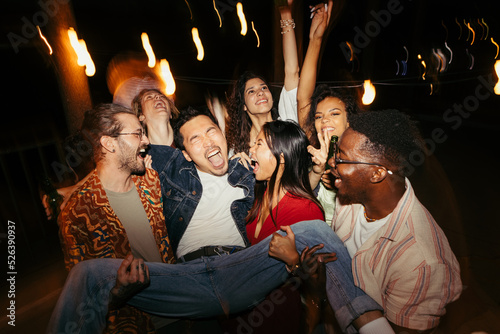 Multiracial friends carry a man at the open-air rooftop night party.