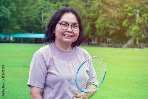 Portrain of asian middle aged woman holding badminton racket, standing and smiling in the green field to play outdoor badminton, concept for to be strong and healthy woman by exercising afterwork.