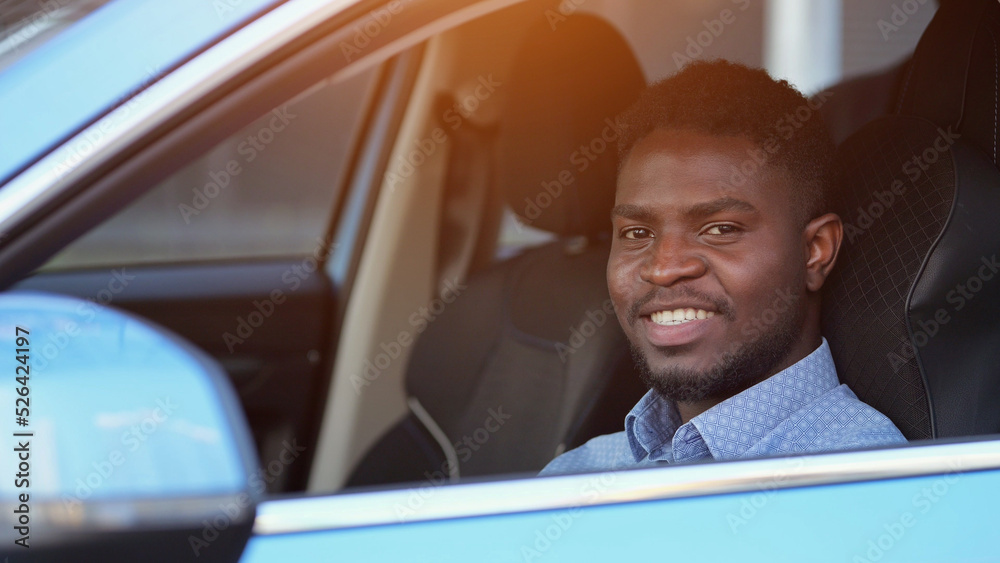 African American driver sits in car dark cabin. Bearded businessman stuck in traffic jam on road driving blue car. Black man enjoys life showing toothy smile, sunlight