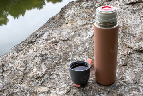 Beige thermos bottle and cup of tea or coffee on rock over the river. Beautiful summer, spring, autumn landscape. Picnic on the rock