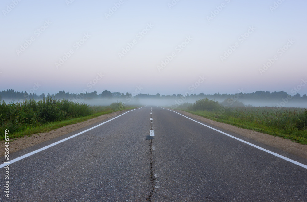 Empty, straight suburban asphalt highway with white road markings vanishing into the morning mist at dawn. Yaroslavl Oblast, Russia.