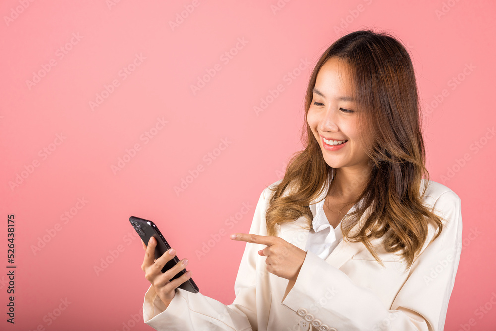 Portrait Asian happy beautiful young woman smile making finger pointing on screen smartphone in hand shopping online application looking to screen, studio shot isolated pink background with copy space Portrait Asian happy beautiful young woman smile making finger pointing on screen smartphone in hand shopping online application looking to screen, studio shot isolated pink background with copy space