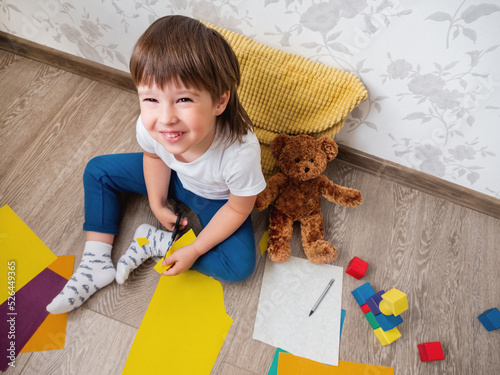 Toddler boy learns to cut colored paper with scissors. Kid sits on floor in kids room with toy blocks and teddy bear. Developing feeling sensations and fine motor skills at home. photo