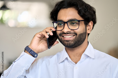 Smiling young indian business man bank manager agent holding smartphone talking consulting client on cell phone making call in office working consulting client providing business service by cellphone.