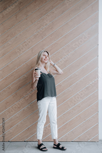 Smiling young lady standing at the wooden wall outdoors and holding a cup of coffee.