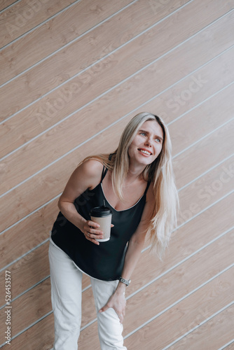 Smiling young lady standing at the wooden wall outdoors and holding a cup of coffee.