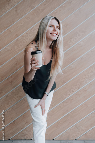 Smiling young lady standing at the wooden wall outdoors and holding a cup of coffee.