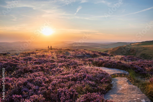Absolutely beautiful sunset landscape image unidentified young couple looking from Higger Tor in Peak District across to Hope Vally in late Summer with heather in full purple bloom